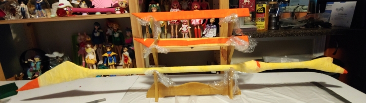The stick and a pair of swords drying on a wooden display rack. The wood is covered with plastic wrap to protect it.