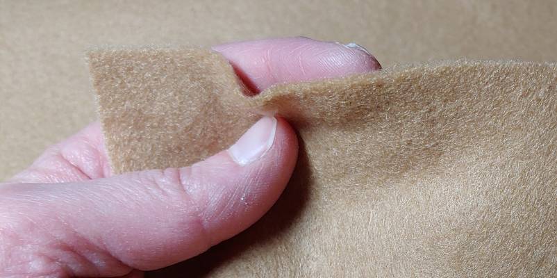 Beige acrylic felt showing off the fibers that make up the fabric pinched between my fingers the usual thickness of a good cut of felt