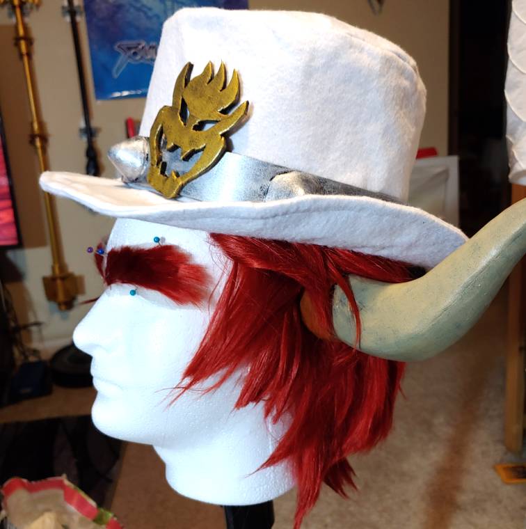 White felt Bowser hat sits on a wig head with red hair and beige horns. The hat has a gold bowser symbol and silver band